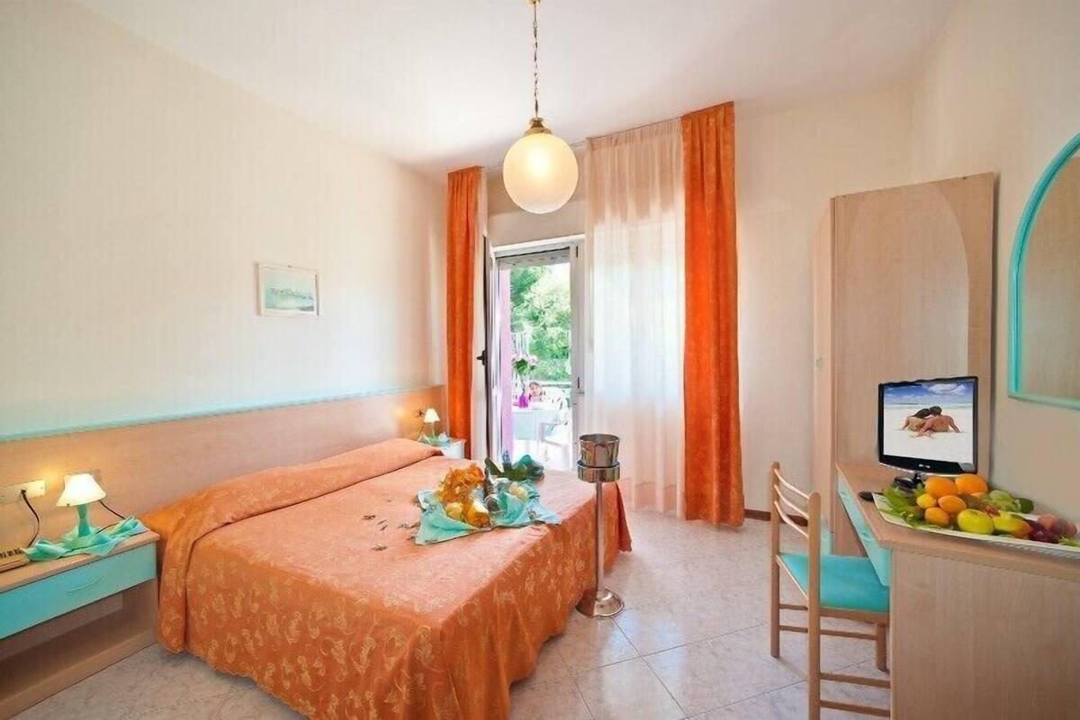 Een kamer met een bed, een bureau met een computer en fruit, en uitzicht op de natuur.