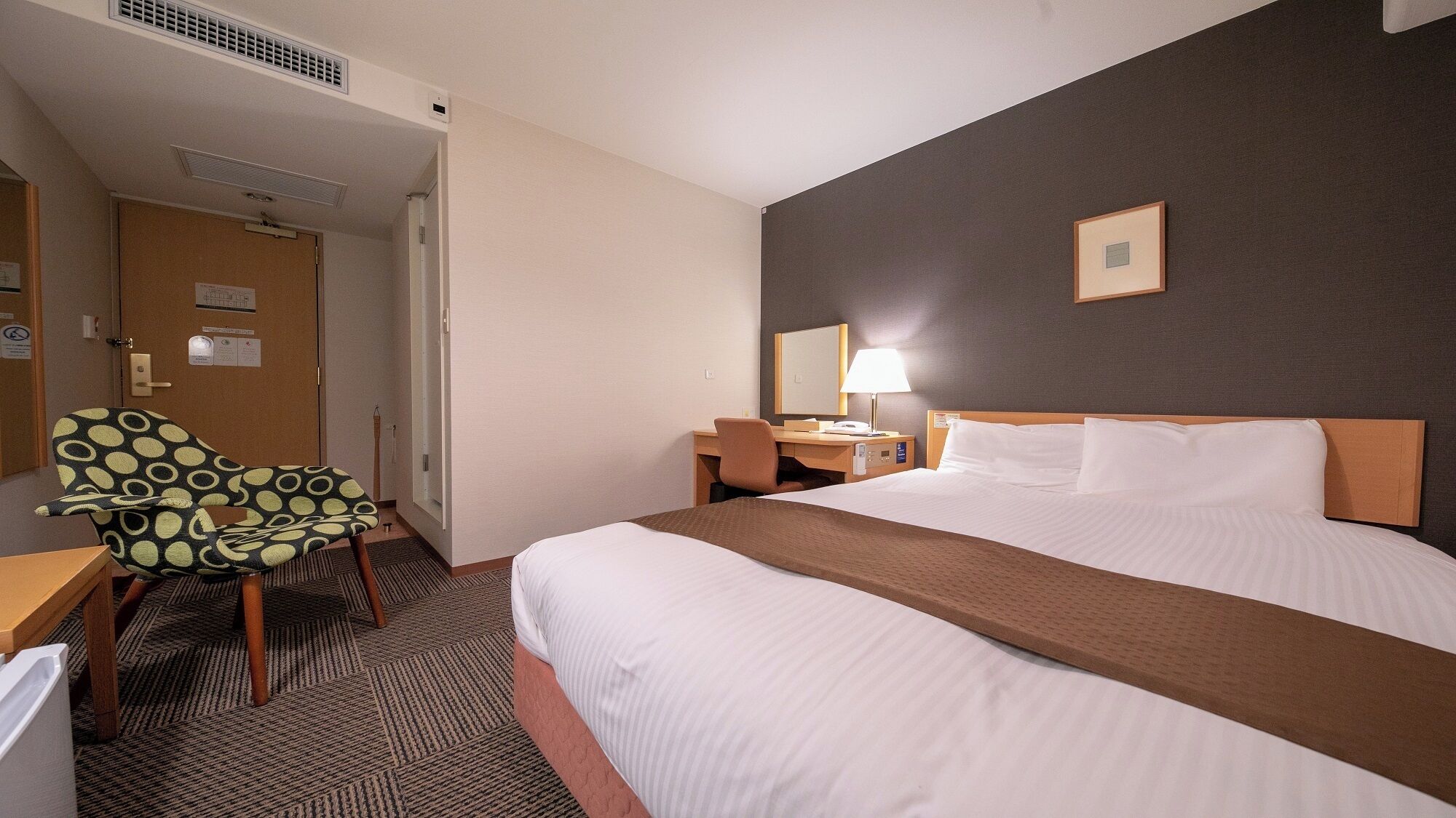 A hotel room with a bed, a desk, a chair, a bedside table, a lamp, and a door.