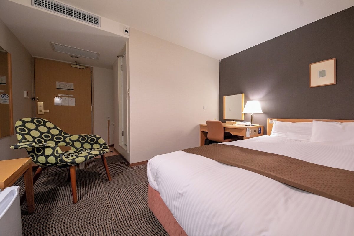 Habitación de hotel con cama, escritorio, silla, mesita de noche, lámpara y puerta.