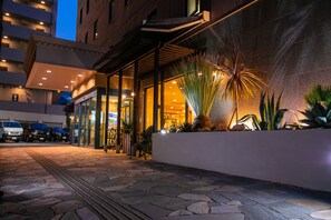 Entrada de hotel con un paseo adoquinado de piedra, una zona de estar cubierta y una exhibición de plantas de palma.