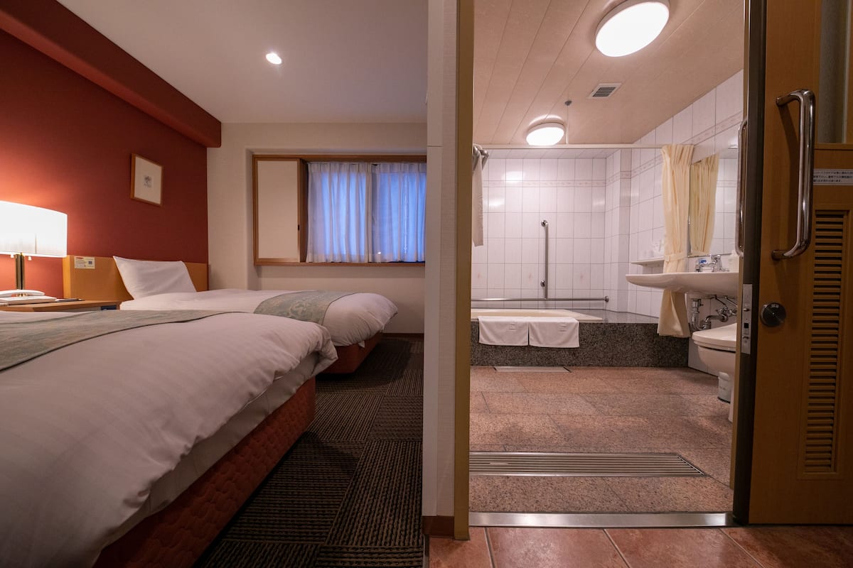 Habitación de hotel con dos camas, una ventana con cortinas, un baño con lavamanos y espejo, y una puerta con manija.