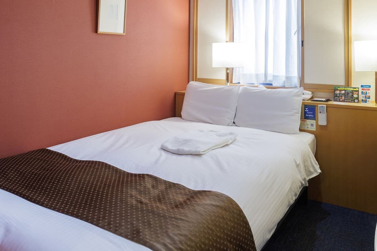 Habitación de hotel con cama, almohadas, mesita de noche con lámpara, televisión y ventana con cortinas.