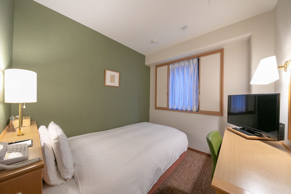 Habitación de hotel con cama, mesita de noche con lámpara, escritorio con televisor y ventana con cortinas.
