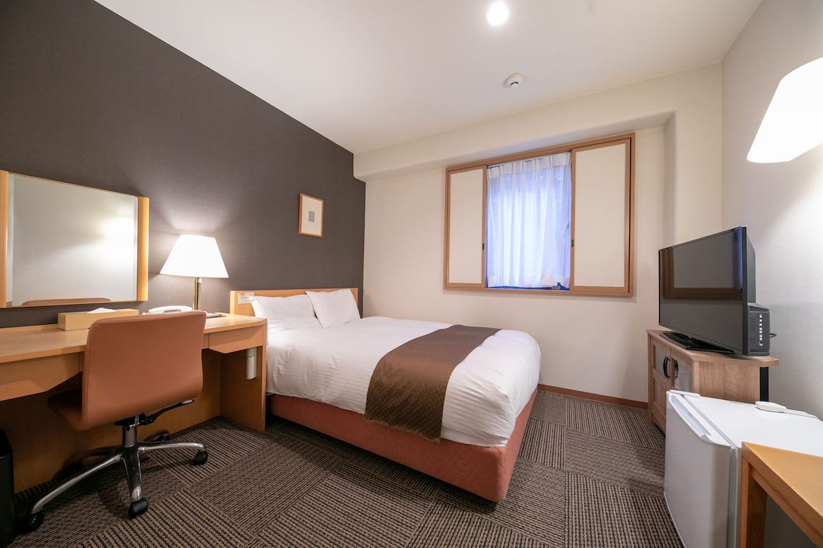 Una habitación de hotel con cama, escritorio, silla, televisor y ventana con cortinas.