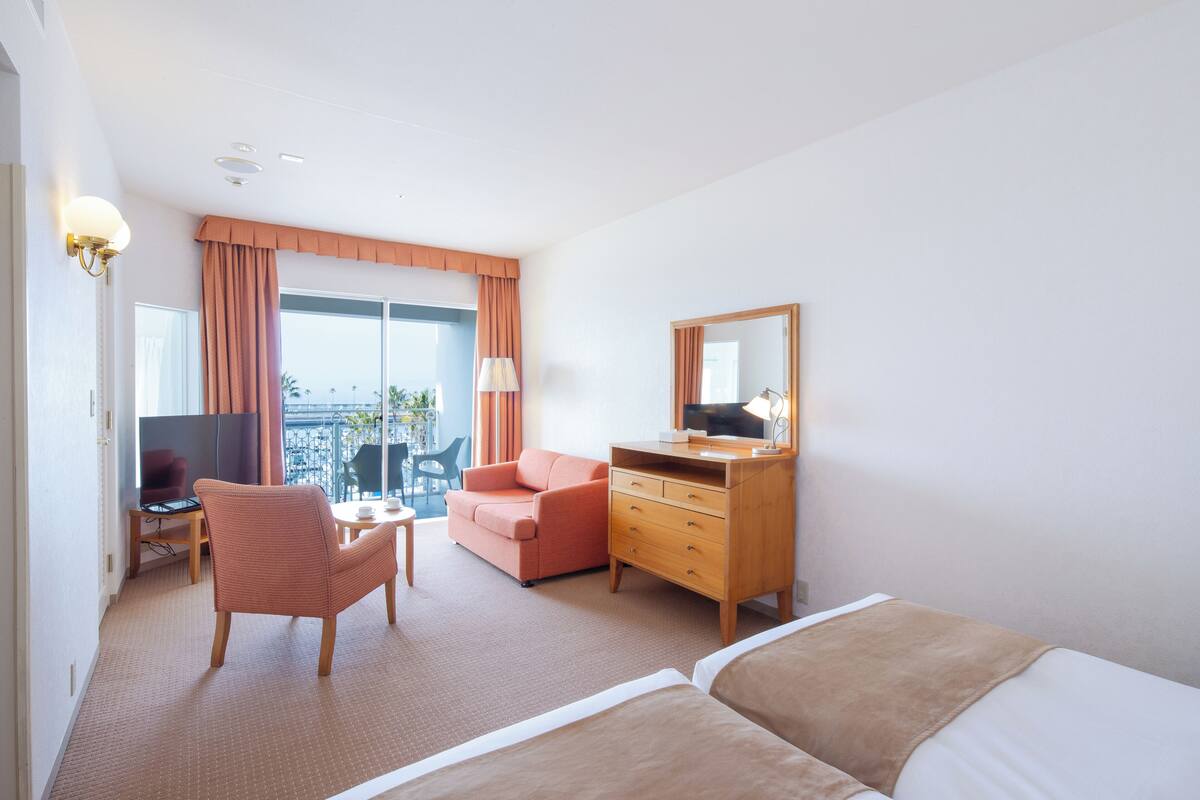 Une chambre d’hôtel avec un lit, un bureau avec une télévision, une chaise, un canapé, une commode et un balcon avec vue.