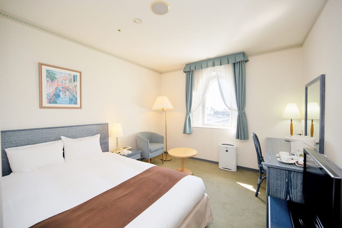 Une chambre d’hôtel comprenant un lit, un bureau, une chaise, une lampe, une petite table et une fenêtre avec des rideaux.