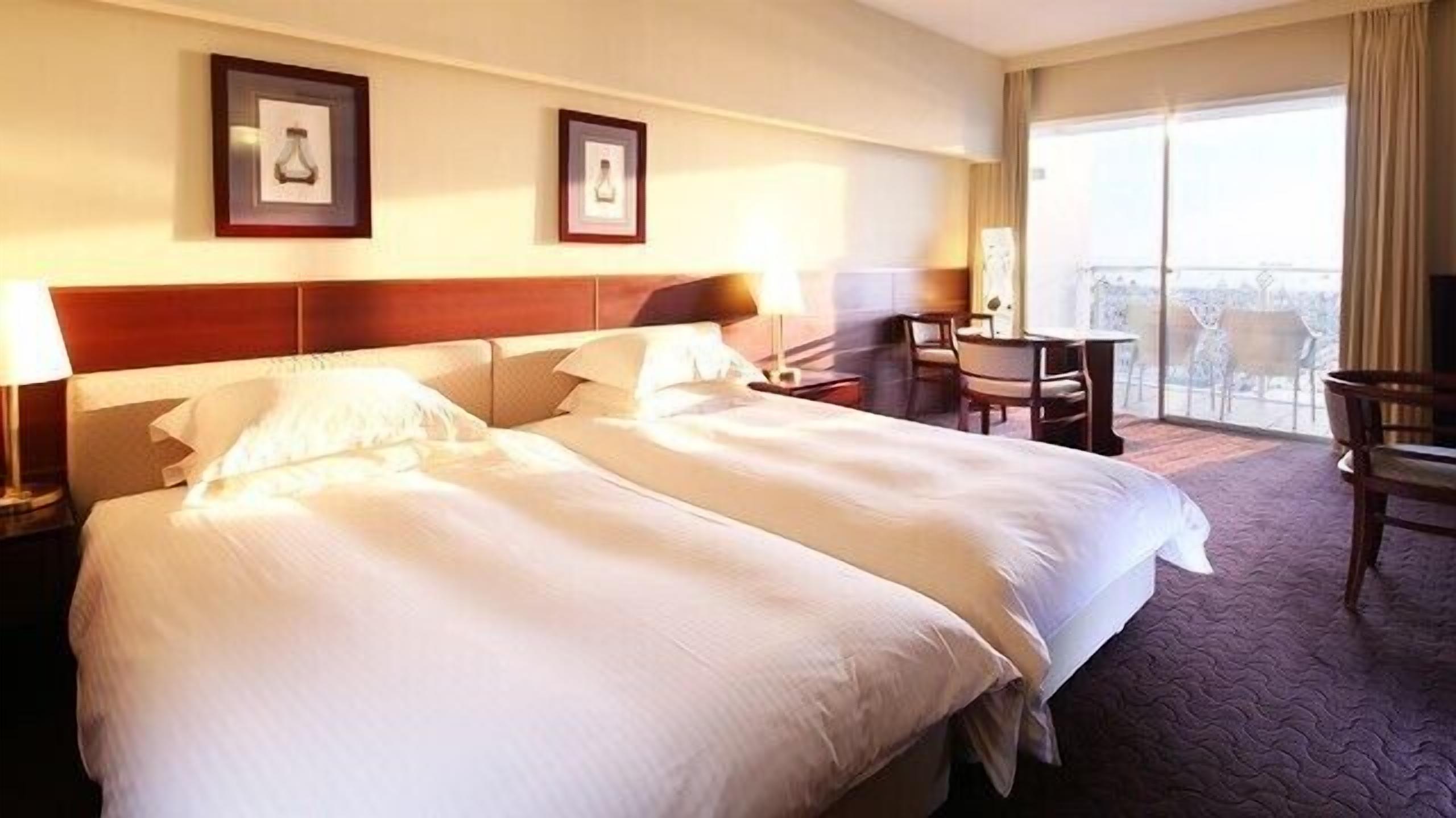 A hotel room with two beds, a desk, chairs, and a balcony with a view.