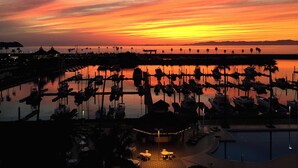 A marina at sunset with boats docked, a pool area with lounge chairs, and a distant view of the ocean and mountains.