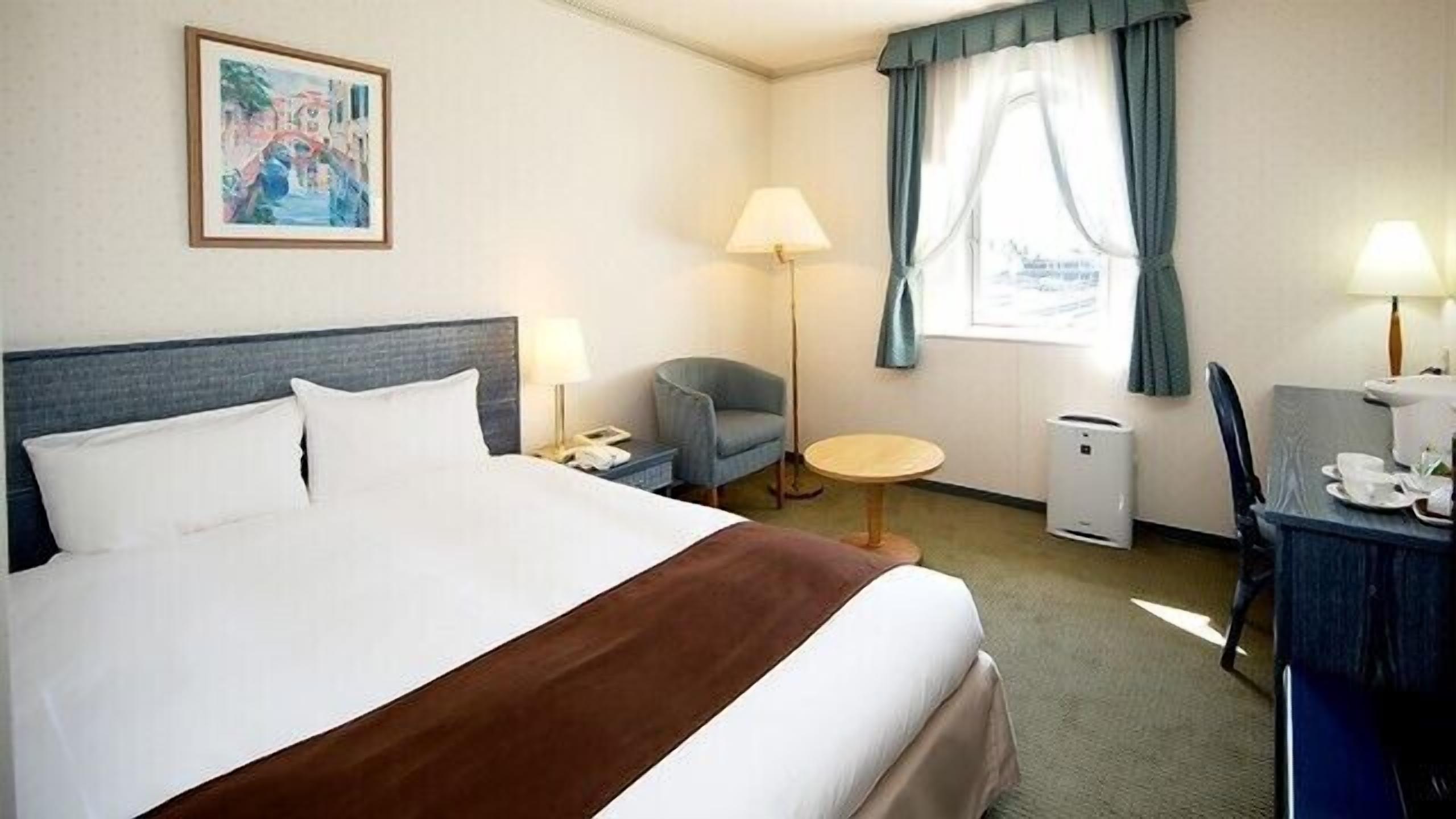 A hotel room with a bed, a chair, a desk, a lamp, a picture on the wall, and a window with curtains.