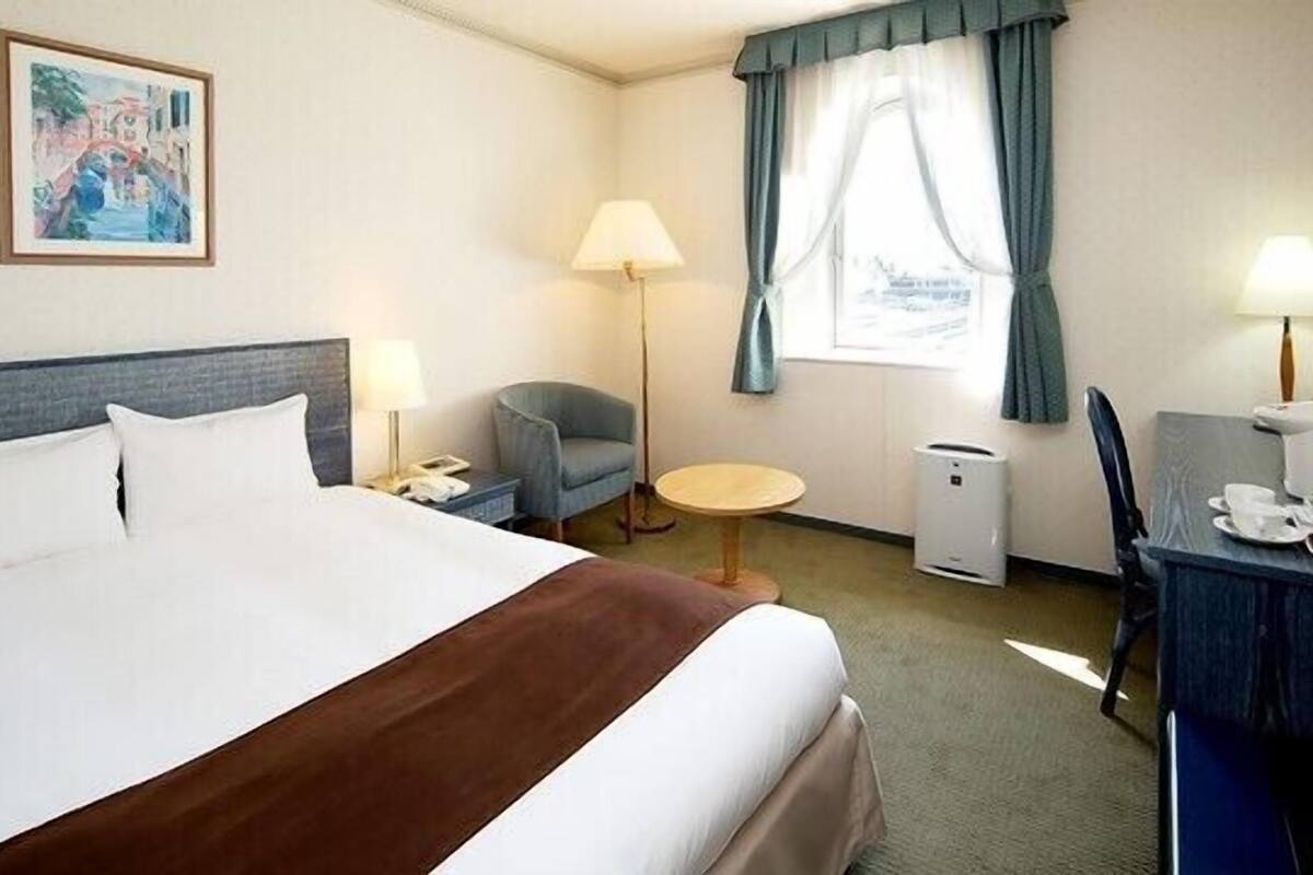 Une chambre d’hôtel avec un lit, une chaise, un bureau, une lampe, un tableau au mur et une fenêtre avec des rideaux.