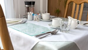 A neatly set dining table with a white tablecloth, a glass placemat, cutlery, a cup, a glass, and a jug.