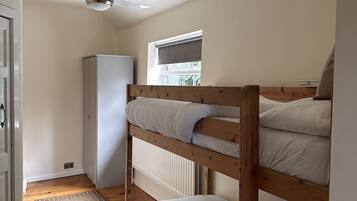 Un dormitorio con literas, ventilador de techo, ventana con persianas, radiador y armario blanco.