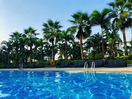 Uma piscina com água azul cristalina, rodeada por palmeiras e uma área de estar com cadeiras.