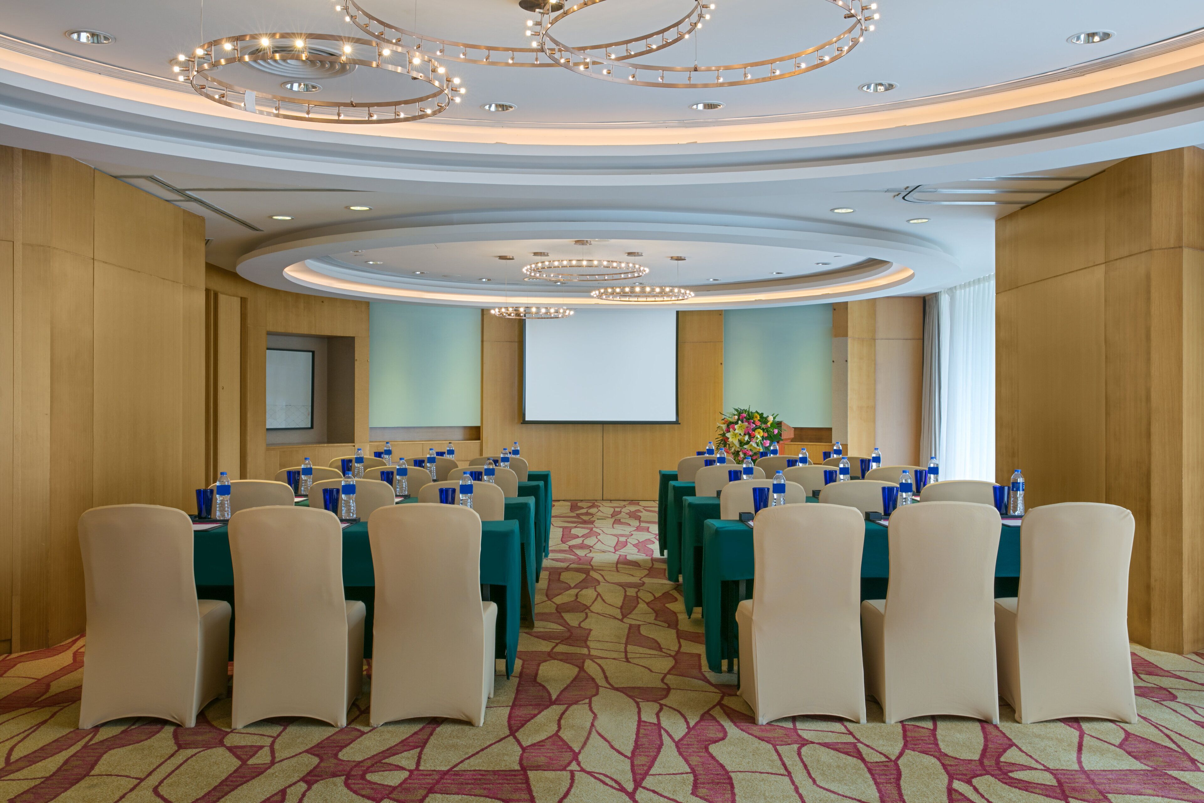 A conference room with rows of tables and chairs, a projector screen, and a floral arrangement.