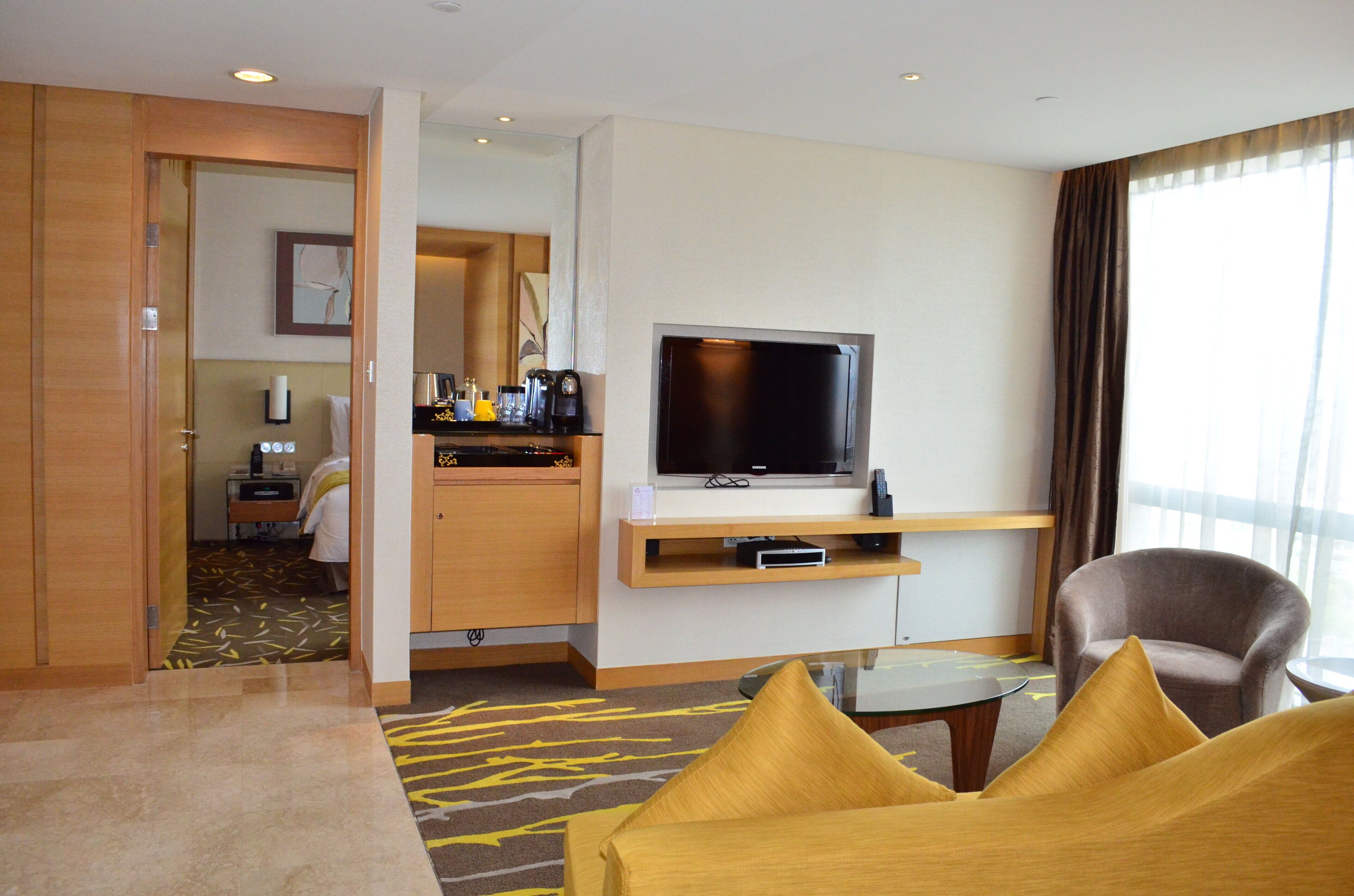 Una habitación de hotel con televisión, sofá, sillón, una mesita y un minibar.