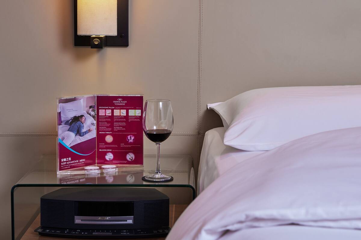 Une chambre d’hôtel avec un lit parfaitement fait, une table de chevet sur laquelle se trouve un verre de vin rouge, une brochure et un appareil électronique noir.