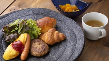 Um prato com alimentos, incluindo um croissant, uma salsicha, uma salada, uma cesta de frutas e uma xícara de café, sobre uma mesa de madeira.