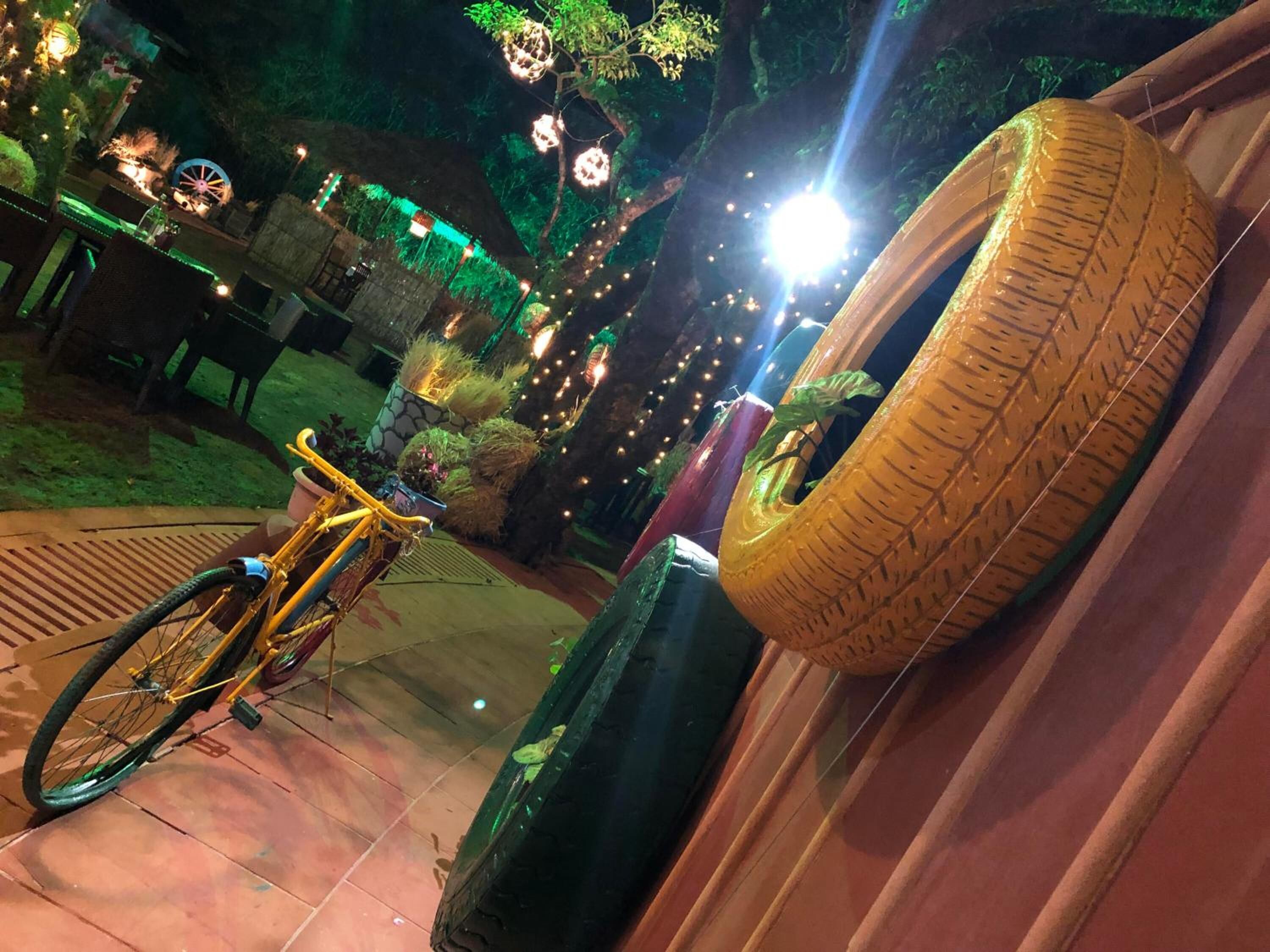 A bicycle parked next to a tire on a tiled patio with string lights and greenery in the background.