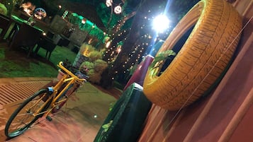 A bicycle parked next to a tire on a tiled patio with string lights and greenery in the background.