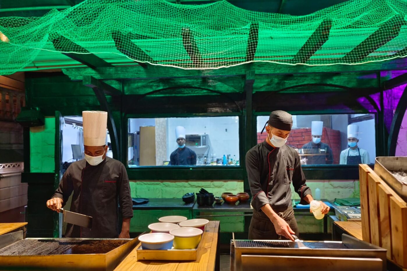 A kitchen with chefs preparing food, wooden tables, and a green net overhead.