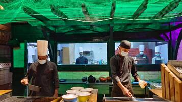 A kitchen with chefs preparing food, wooden tables, and a green net overhead.
