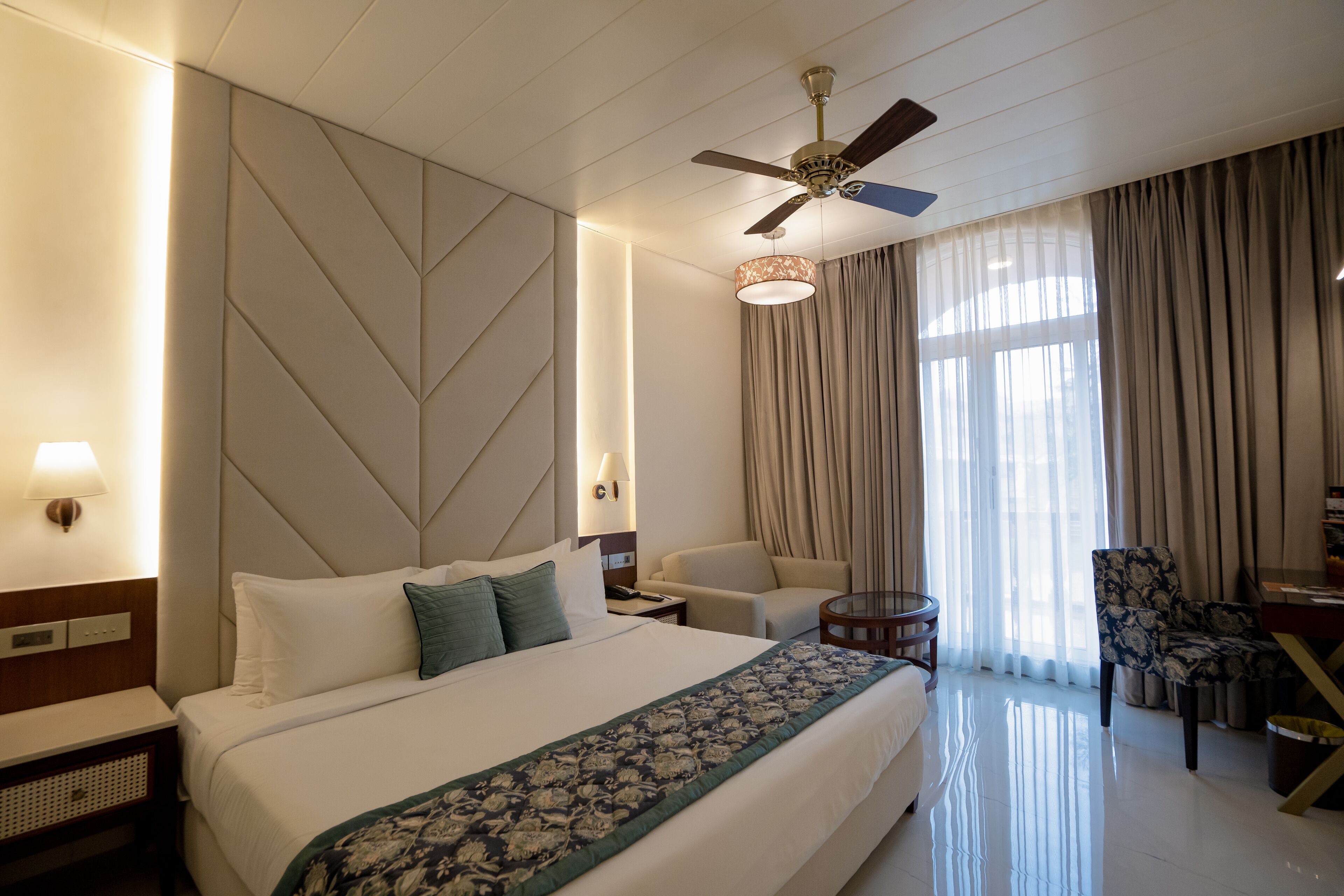 A hotel room with a large bed, a ceiling fan, a desk with a chair, and a window with curtains.
