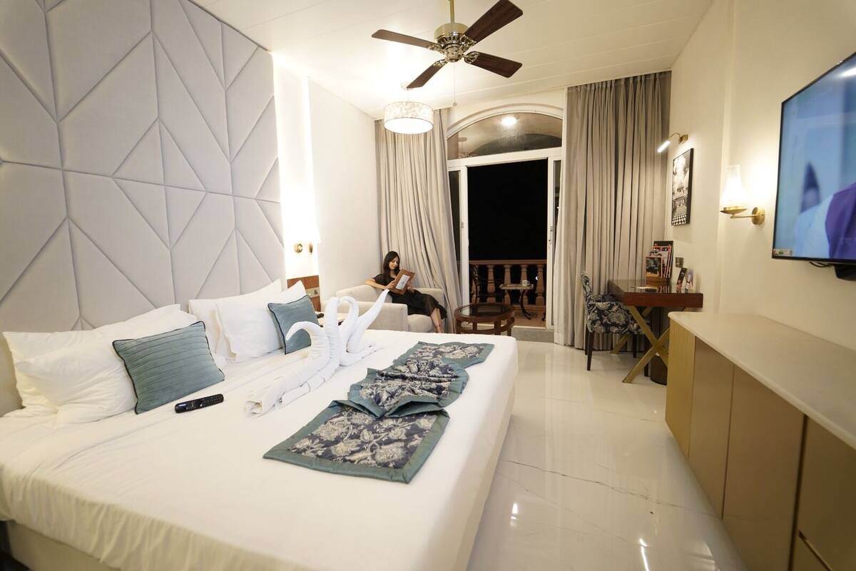 Habitación de hotel con una cama grande, ventilador de techo, televisor y balcón con vistas.