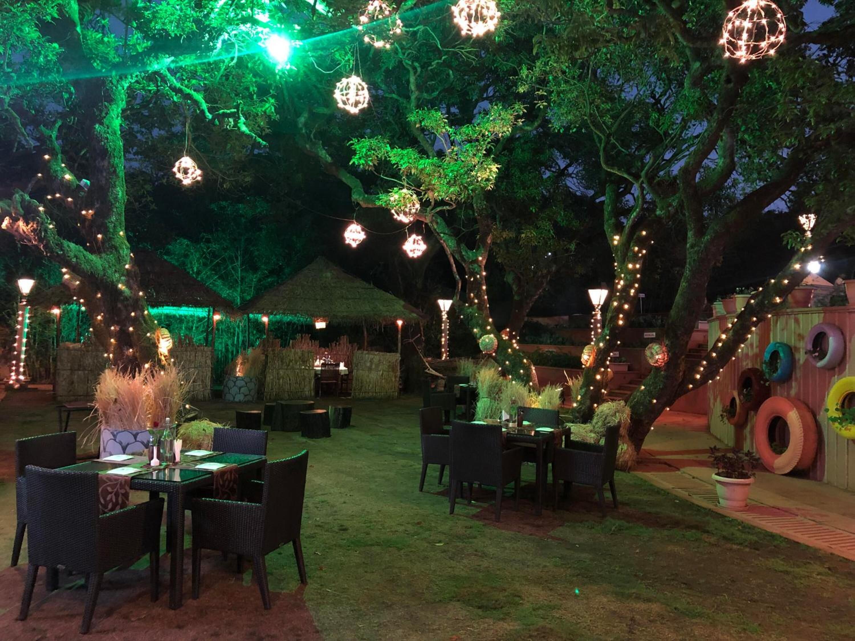 An outdoor dining area with tables and chairs, surrounded by trees and string lights.