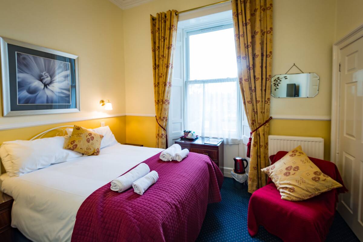 A hotel room with a bed, a chair, a desk, a window with curtains, a radiator, and a mirror on the wall.