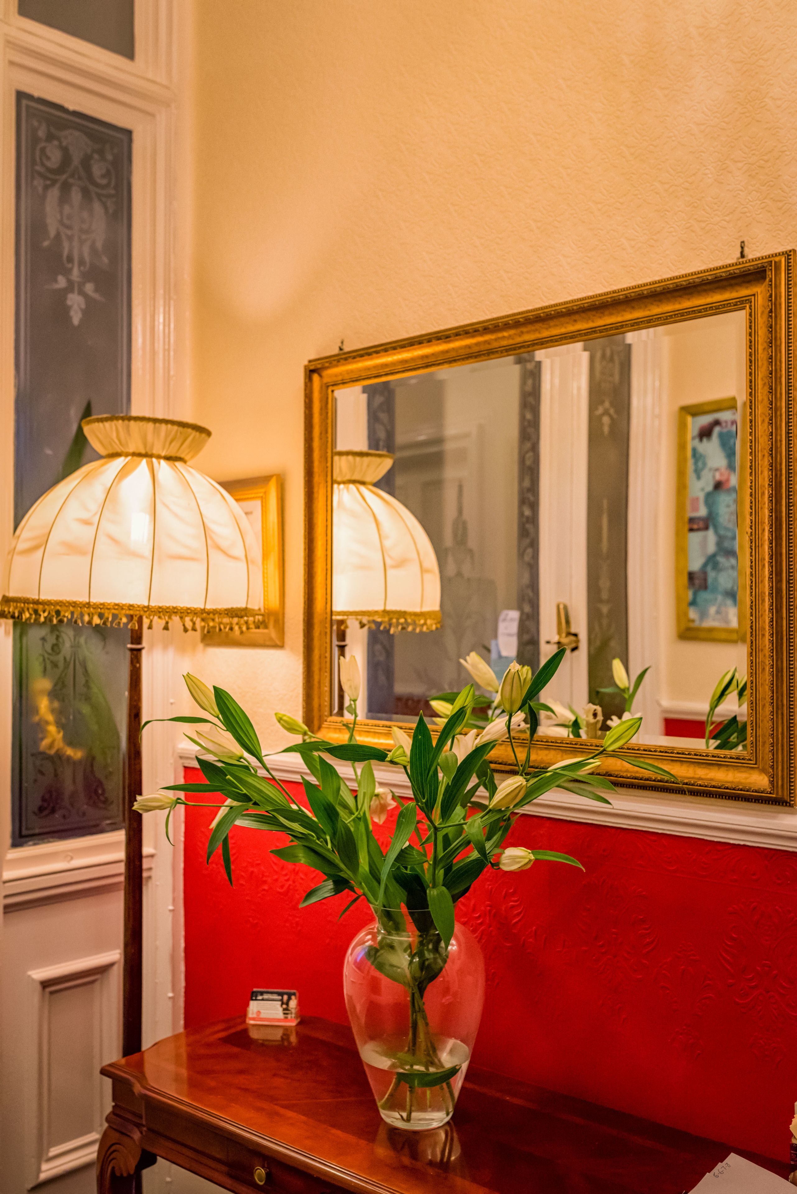 A room with a red wall, a large mirror with a gold frame, and two lamps on a wooden table.