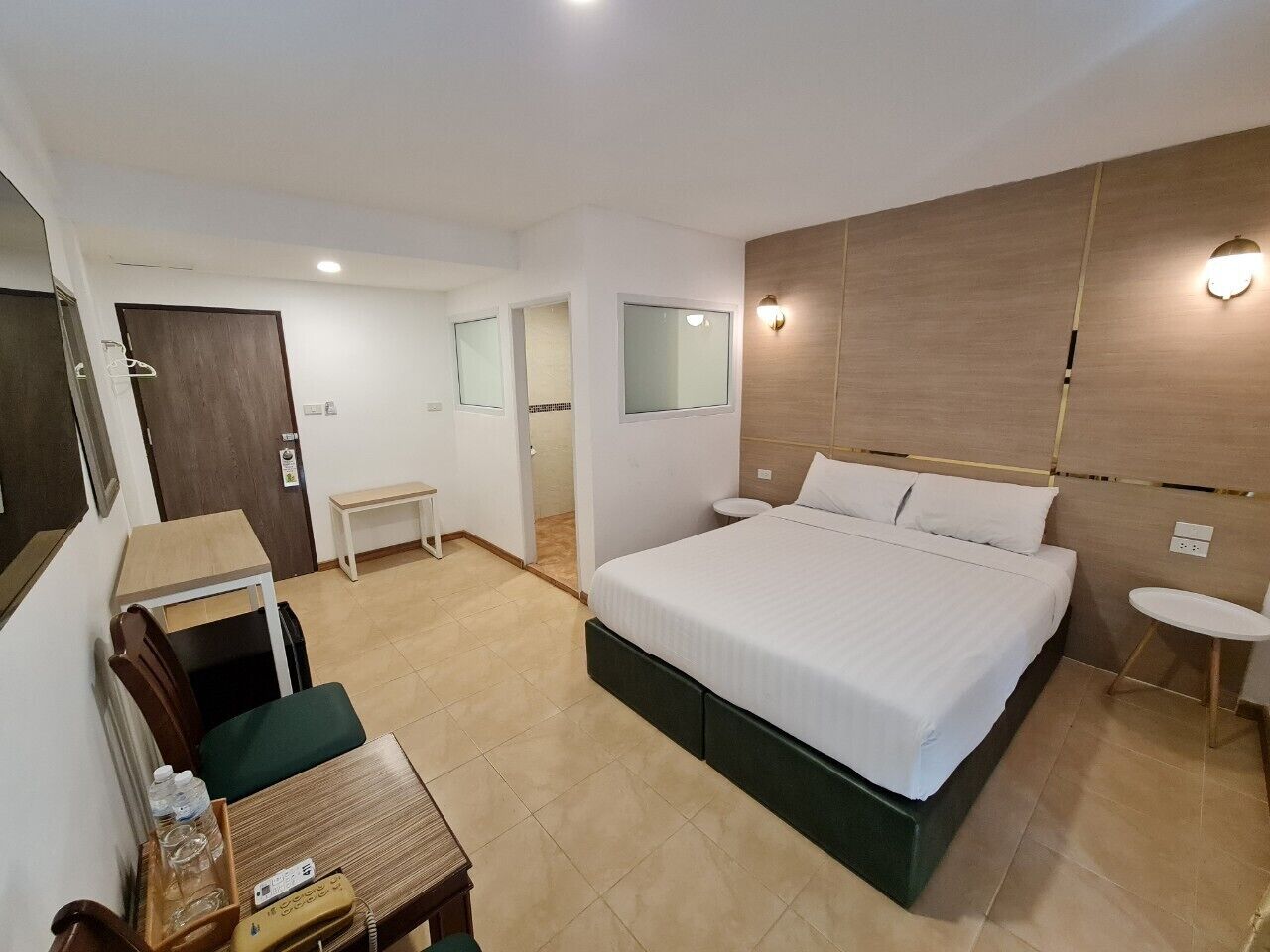 A hotel room with a bed, a desk, a chair, a small table, a bench, and a wall-mounted light fixture.