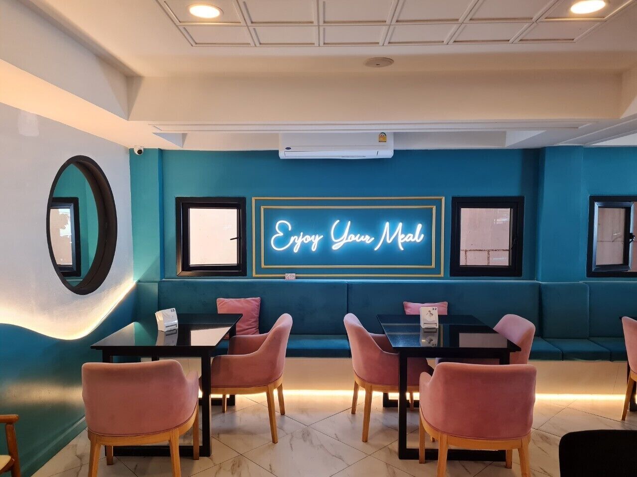 Interior de restaurant amb parets color turquesa, un cartell de neó que diu "Gaudeix del teu àpat" i cadires roses al voltant de taules.