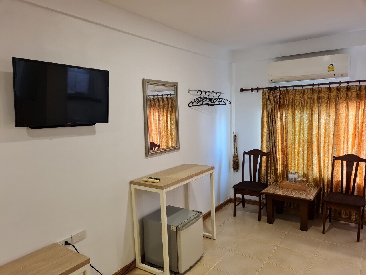 A living room with a flat-screen TV, a small table, a refrigerator, two wooden chairs, and a window with curtains.