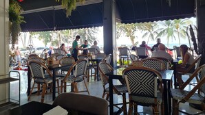 An outdoor restaurant with patterned chairs and tables, a waiter attending to guests, and a view of palm trees and the sea. - Opal House Hotel & Restaurant (Pattaya)