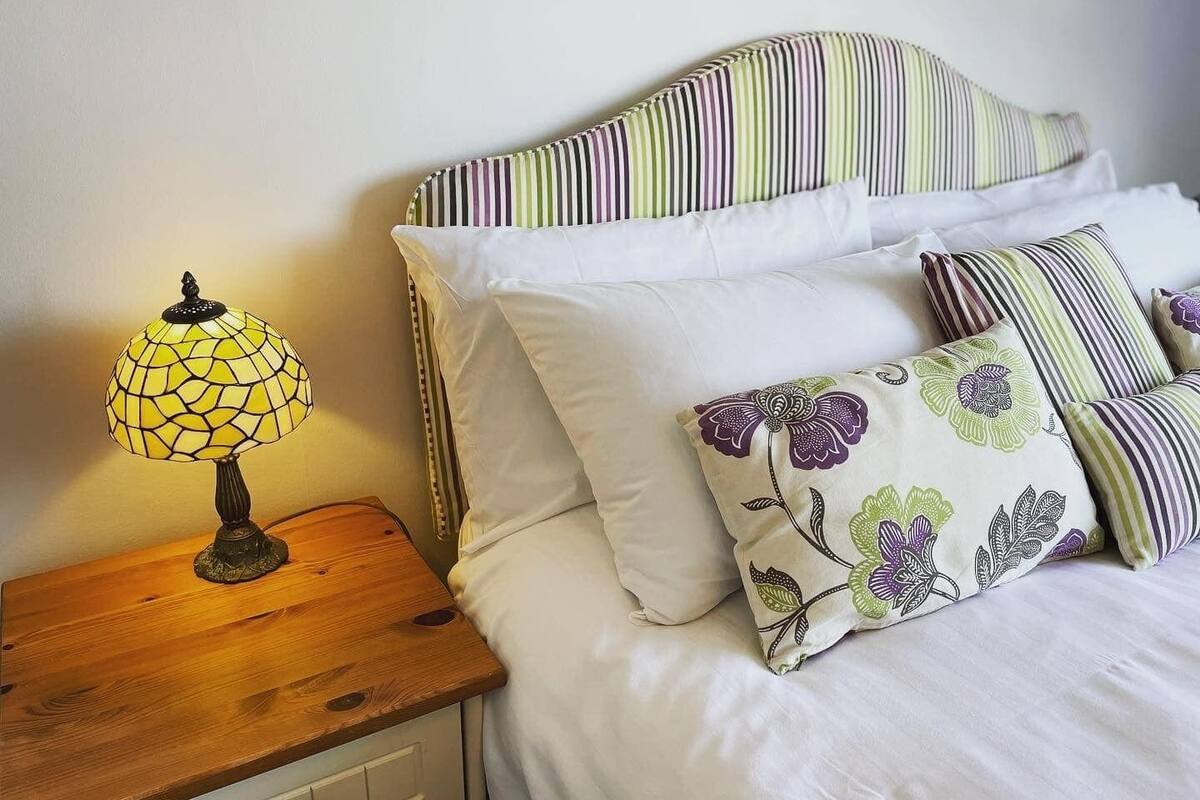 A neatly made bed with striped bedding and floral-patterned pillows, a wooden nightstand with a lamp, and a plain wall.