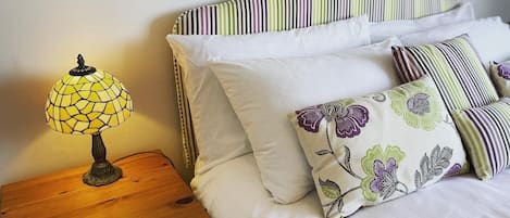 A neatly made bed with striped bedding and floral-patterned pillows, a wooden nightstand with a lamp, and a plain wall.
