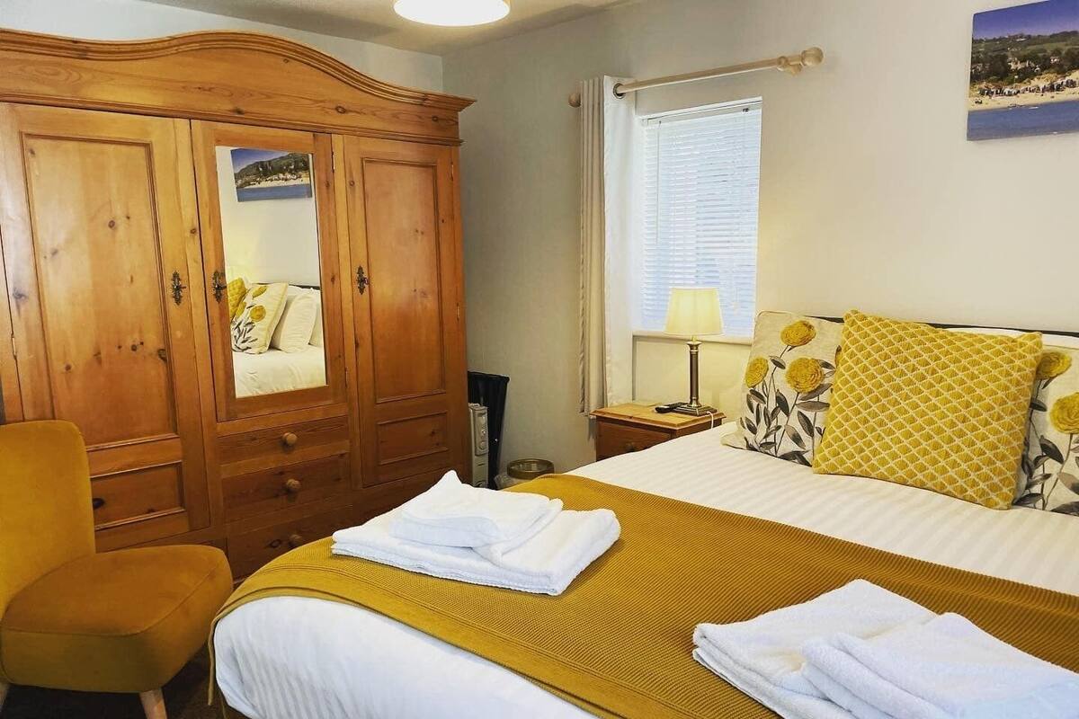 A hotel room with a bed, a wooden wardrobe, a chair, a window with blinds, and a lamp.