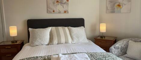 A neatly made bed with white bedding and a green and white blanket, two bedside lamps, and two wall-mounted flower paintings.