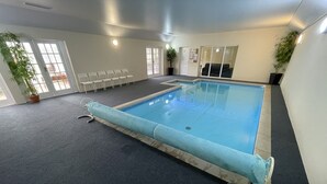 Una piscina interior con superficie de agua azul, rodeada por un suelo oscuro alfombrado, sillas blancas y plantas en macetas.