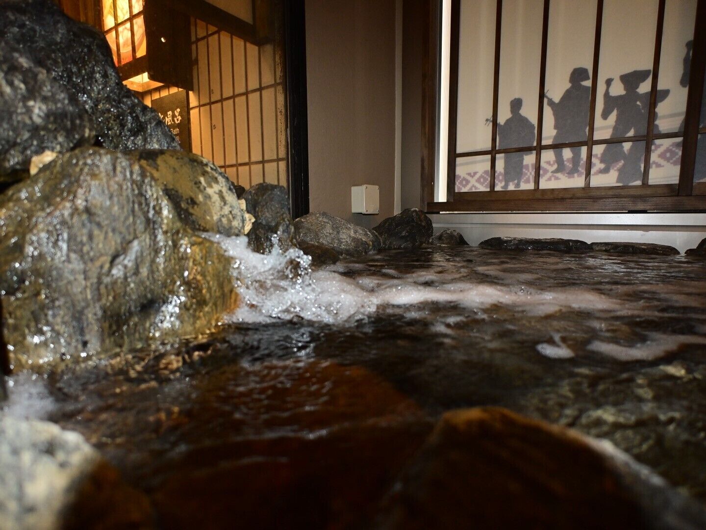 Una fuente de piedra con un arroyo, una habitación tradicional japonesa con suelo de tatami y una vista a través de un celosía shoji.