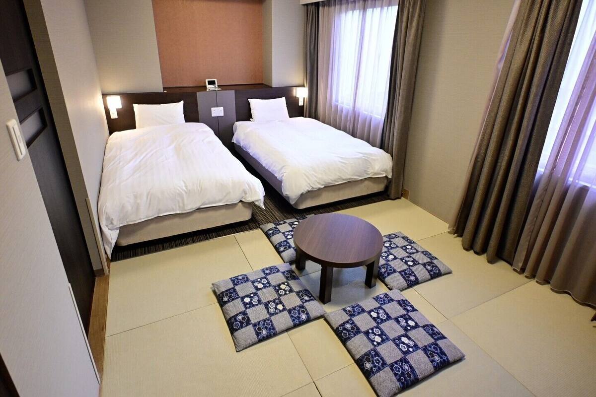 A hotel room with two beds, a small table, and a view of the outside through the window.