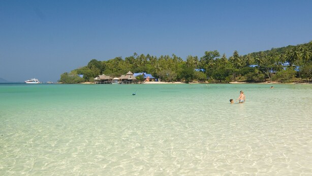 A beach with clear turquoise water, a person in the water, and a row of buildings with thatched roofs in the background. - Koh Talu Island Resort (Bang Saphan Noi)