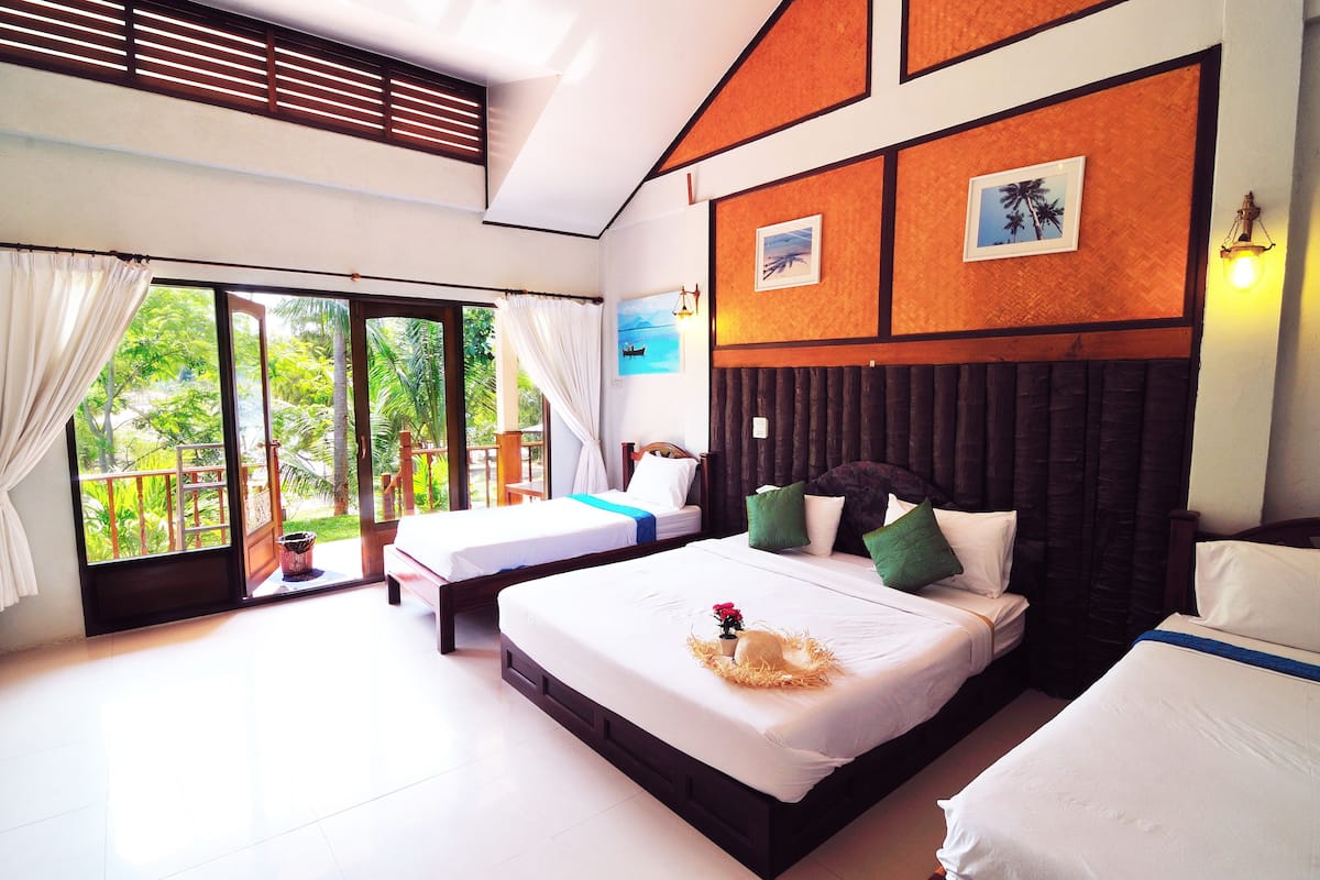 A hotel room with two beds, a balcony with a view of greenery, and a wooden headboard.