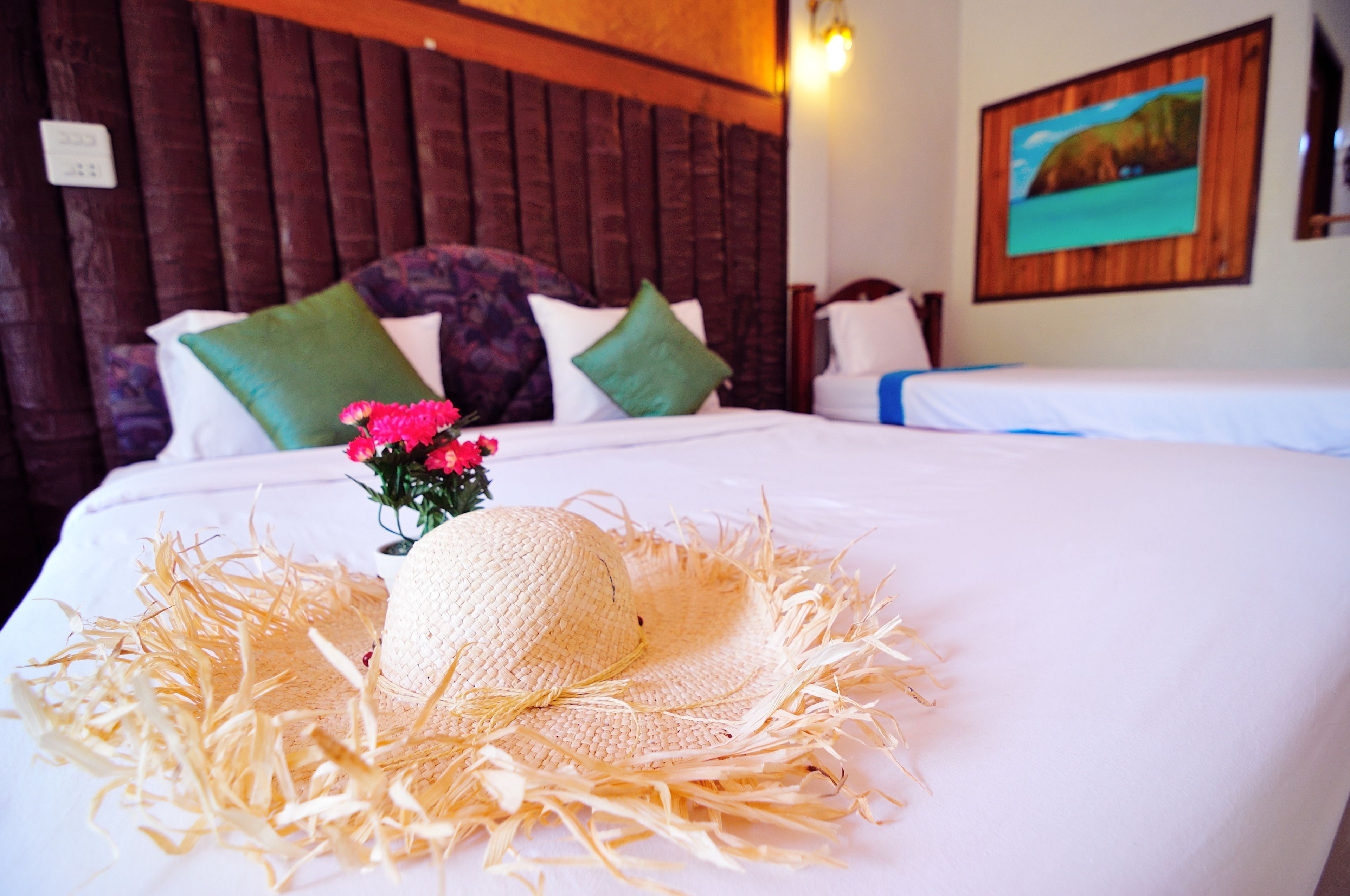 Una cama bien hecha con un sombrero de paja y flores, cabecera de madera y un cuadro en la pared.