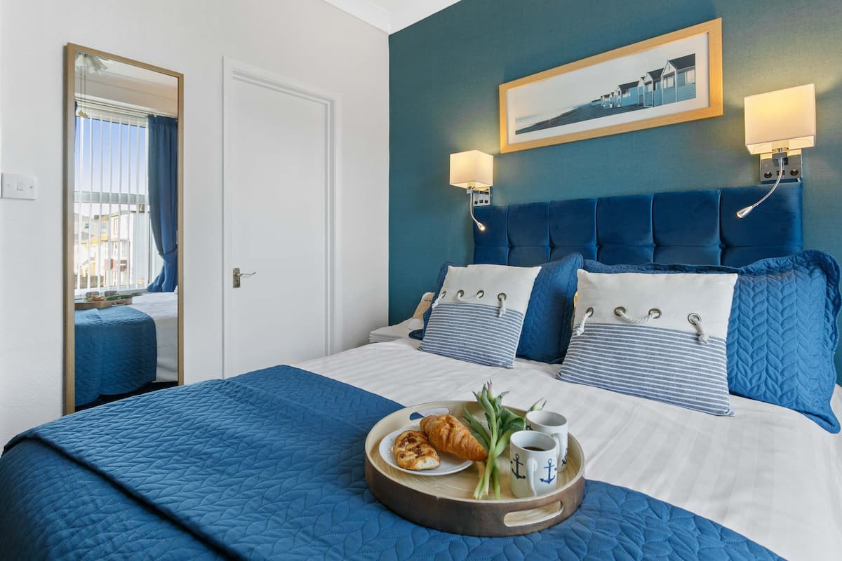 A hotel room with a large bed, a headboard with lights, a framed picture on the wall, and a tray with croissants and coffee on the bed.