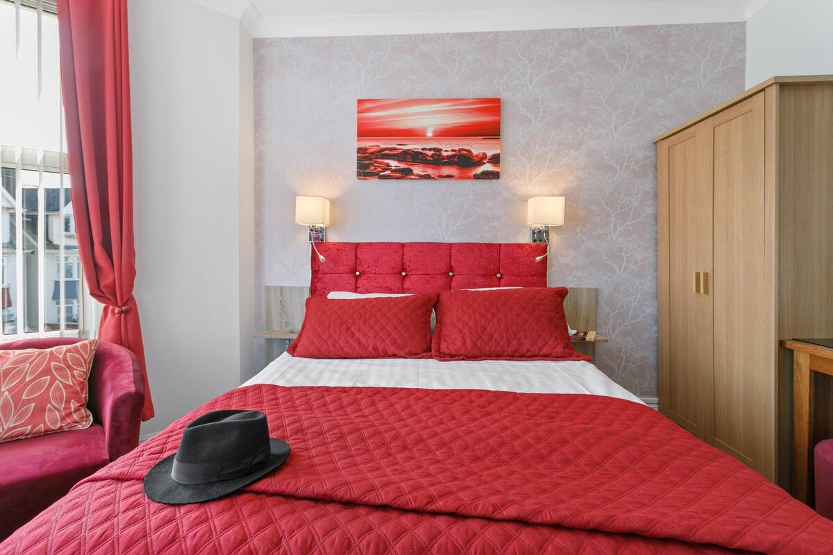 A hotel room with a red bedspread, a black hat on the bed, a red upholstered chair, a wooden wardrobe, and a wall-mounted painting.
