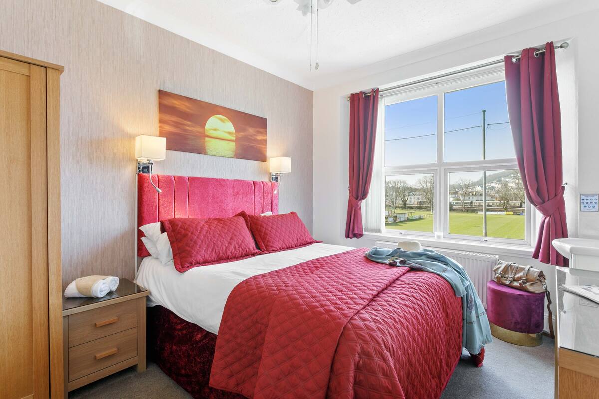 A hotel room with a large bed, a nightstand, a window with curtains, and a view of the outside.