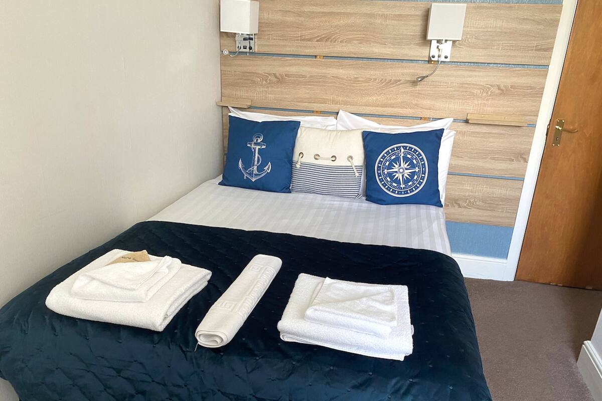 A neatly made bed with a dark quilt, two rolled towels, and two pillows with nautical designs.