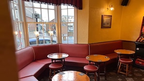 A corner seating area with red cushioned benches and round tables, a framed picture on the wall, and a view of the street outside. - The Brunswick Hotel (Bridlington)