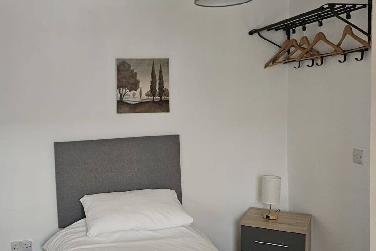 A neatly made bed with white linens and towels, a grey headboard, a wall-mounted lamp, a small wooden nightstand with drawers, and a simple painting of trees on the wall.